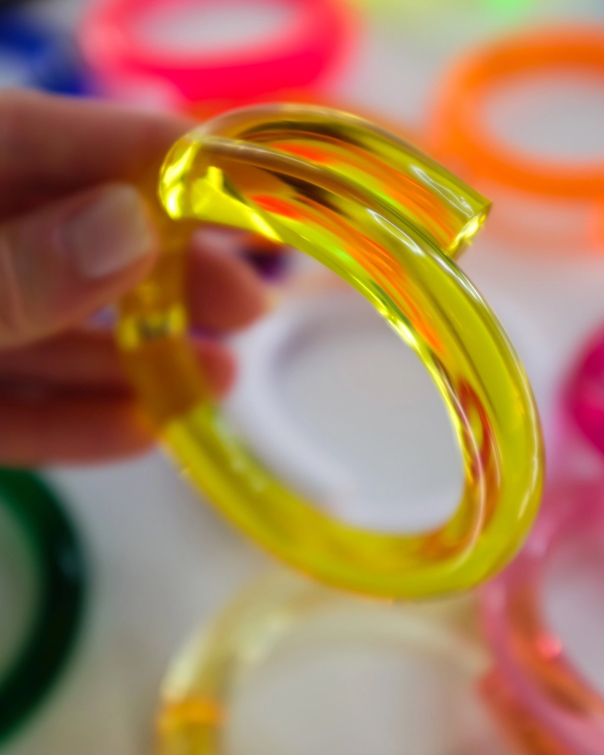 A hand holds the Faire Lucite Yellow Neon Bracelet, with other colorful handcrafted acrylic bangles in the background—ideal for minimalist jewelry lovers.