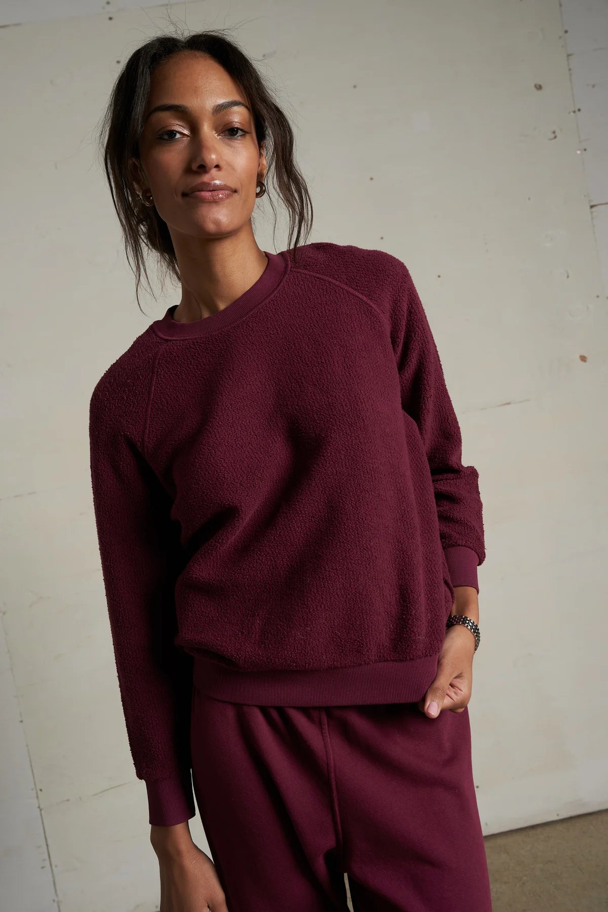 A woman stands indoors wearing the Perfectwhitetee Ziggy Reverse LS Sweatshirt and matching pants in textured maroon cotton fleece, gazing calmly at the camera against a plain light background.