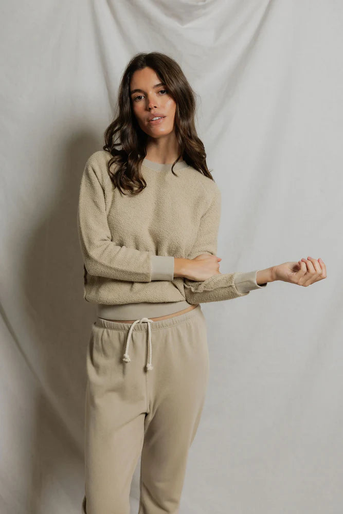 A woman with long brown hair wears the Perfectwhitetee Ziggy Reverse LS Sweatshirt in beige with matching drawstring sweatpants, standing before a light fabric backdrop and looking at the camera with a relaxed expression.
