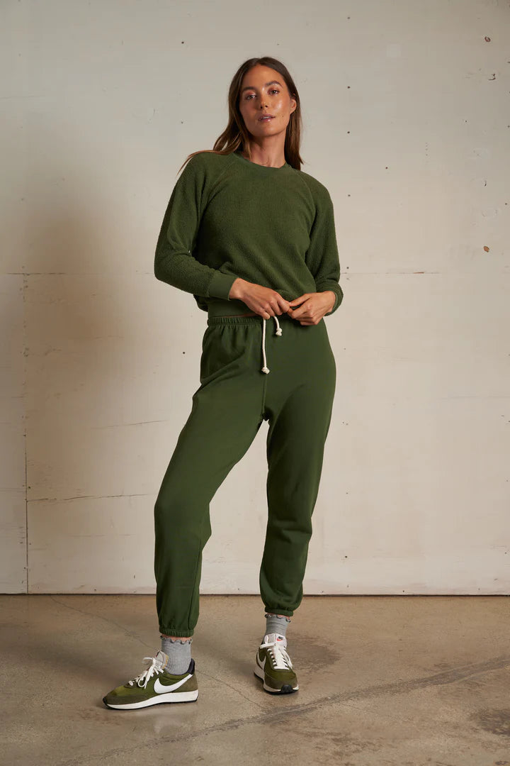 A woman stands indoors against a plain wall, wearing the Perfectwhitetee Ziggy Reverse LS Sweatshirt in olive green with matching jogger pants, gray socks, and green and white Nike sneakers. She looks confidently at the camera, hands on her waist.