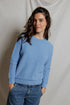 A woman with long brown hair wears the Perfectwhitetee Ziggy Reverse LS Sweatshirt in light blue and blue jeans, standing with hands in pockets and smiling softly in front of a plain, light-colored draped backdrop.