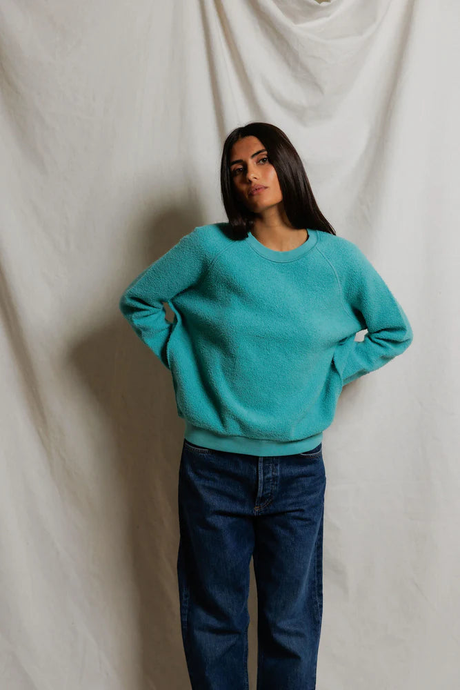 A woman with long dark hair stands before a cream-colored backdrop, confidently posing in blue jeans and the Perfectwhitetee Ziggy Reverse LS Sweatshirt with bracelet-length sleeves.