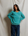 A woman with long dark hair stands before a cream-colored backdrop, confidently posing in blue jeans and the Perfectwhitetee Ziggy Reverse LS Sweatshirt with bracelet-length sleeves.