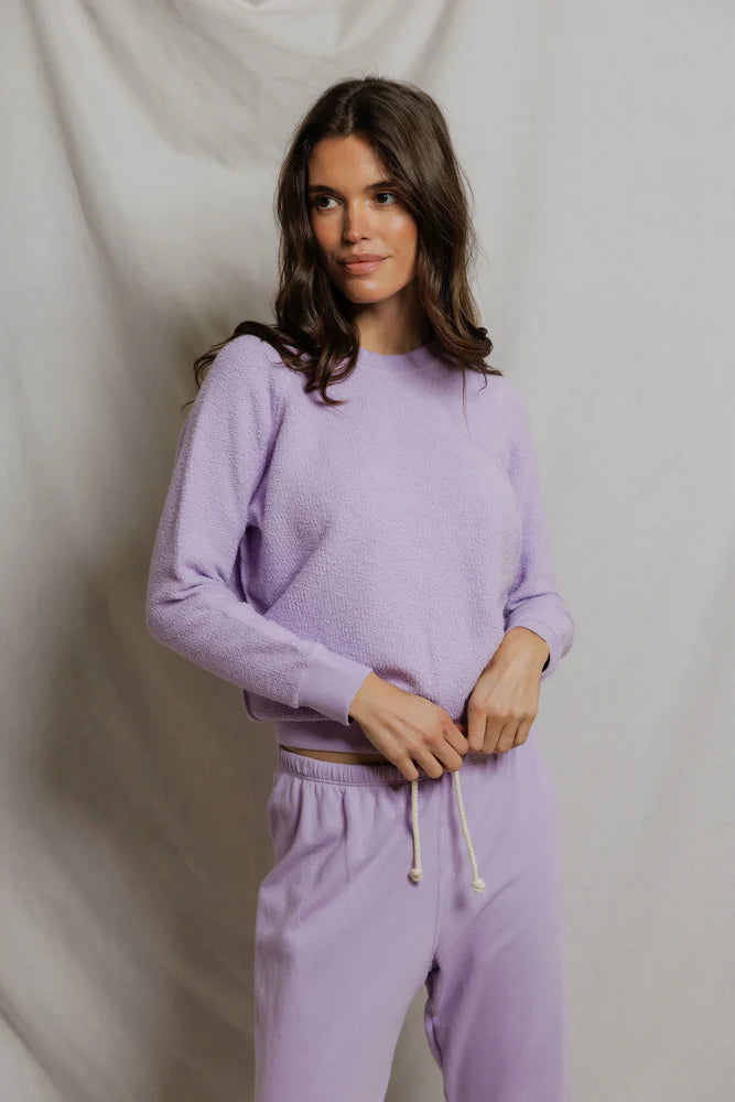 A woman with long brown hair stands in front of a light backdrop, wearing the Perfectwhitetee Ziggy Reverse LS Sweatshirt in light purple with a shrunken fit and bracelet-length sleeves, paired with matching drawstring sweatpants.