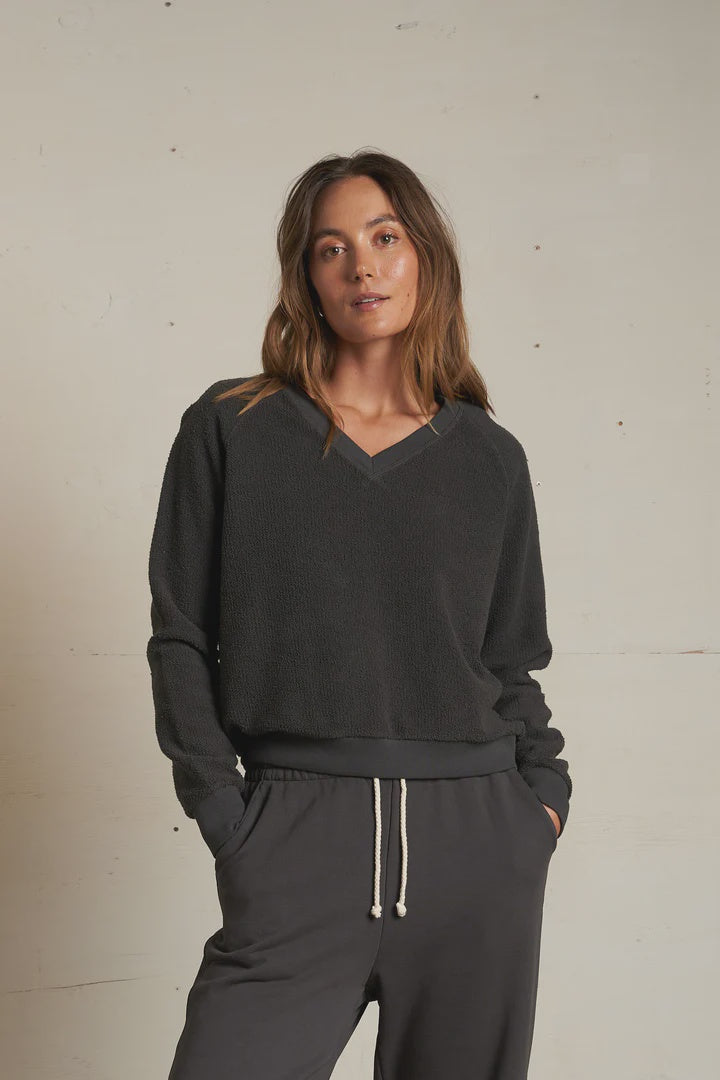 A woman with long brown hair models the Perfectwhitetee Zuri Reverse V Neck, a relaxed dark gray raglan sleeve sweatshirt paired with matching drawstring pants. She stands confidently against a plain light wall, one hand in her pocket.