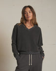 A woman with long brown hair models the Perfectwhitetee Zuri Reverse V Neck, a relaxed dark gray raglan sleeve sweatshirt paired with matching drawstring pants. She stands confidently against a plain light wall, one hand in her pocket.