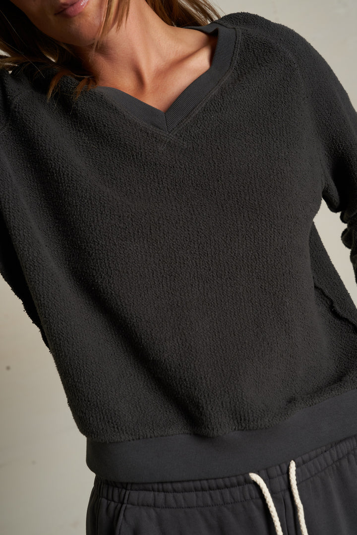Someone wearing the Perfectwhitetee Zuri Reverse V Neck sweatshirt and matching drawstring pants in textured dark gray; the cropped image shows only their midsection, partial face, and relaxed raglan sleeves.