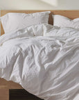 A neatly made bed with Coyuchi Inc's Organic Crinkled Percale Duvet Alpine White Queen and pillows is centered in a bright, minimalist bedroom. A wooden headboard and bedside table with a glass carafe, a tumbler, and a green plant on the side add a touch of warmth and simplicity to the space, showcasing GOTS certified organic cotton bedding.