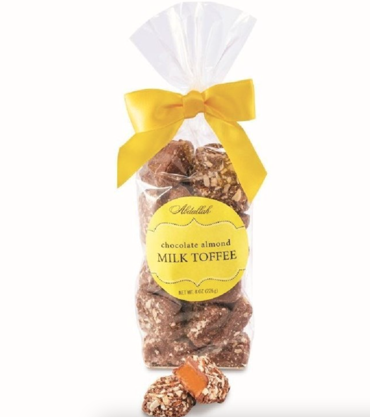 A transparent bag of Abdallah's Butter Almond Toff Mlk is elegantly tied with a yellow ribbon. The label reads "Butter Almond Toff Mlk." Pieces are visible inside the bag, with a few temptingly placed in front.