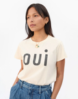 A person with long dark hair wearing a light-colored Classic Tee - OUI by Clare Vivier made in L.A. with the word "oui" written in large black letters across the front. They are also wearing blue high-waisted jeans and are accessorized with necklaces. The 100% cotton T-shirt stands out against the plain white background.