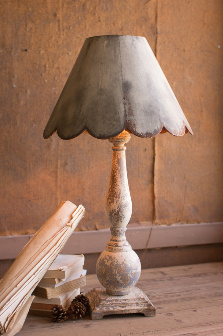 A vintage Metal table lamp with Scalloped Shade by Kalalou, Inc, with a distressed base and a dark gray scalloped lampshade, positioned beside a stack of old books and pine cones on a wooden surface in a Scottsdale, Arizona bungalow against a textured wall.