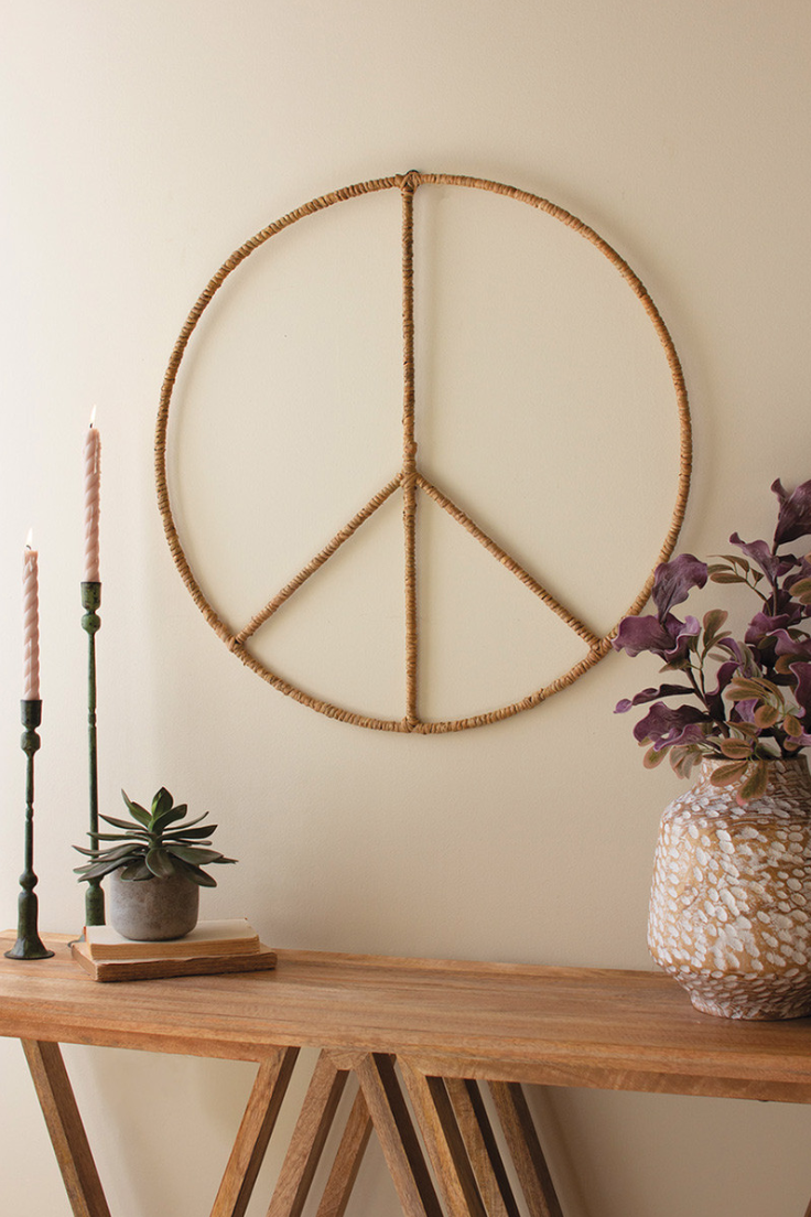 A Seagrass Iron Wall Hanging Peace Sign made by Kalalou, Inc hangs on a beige wall in a Scottsdale, Arizona bungalow, next to a wooden shelf holding a vase with flowers, two pink candles, and a small potted succulent.