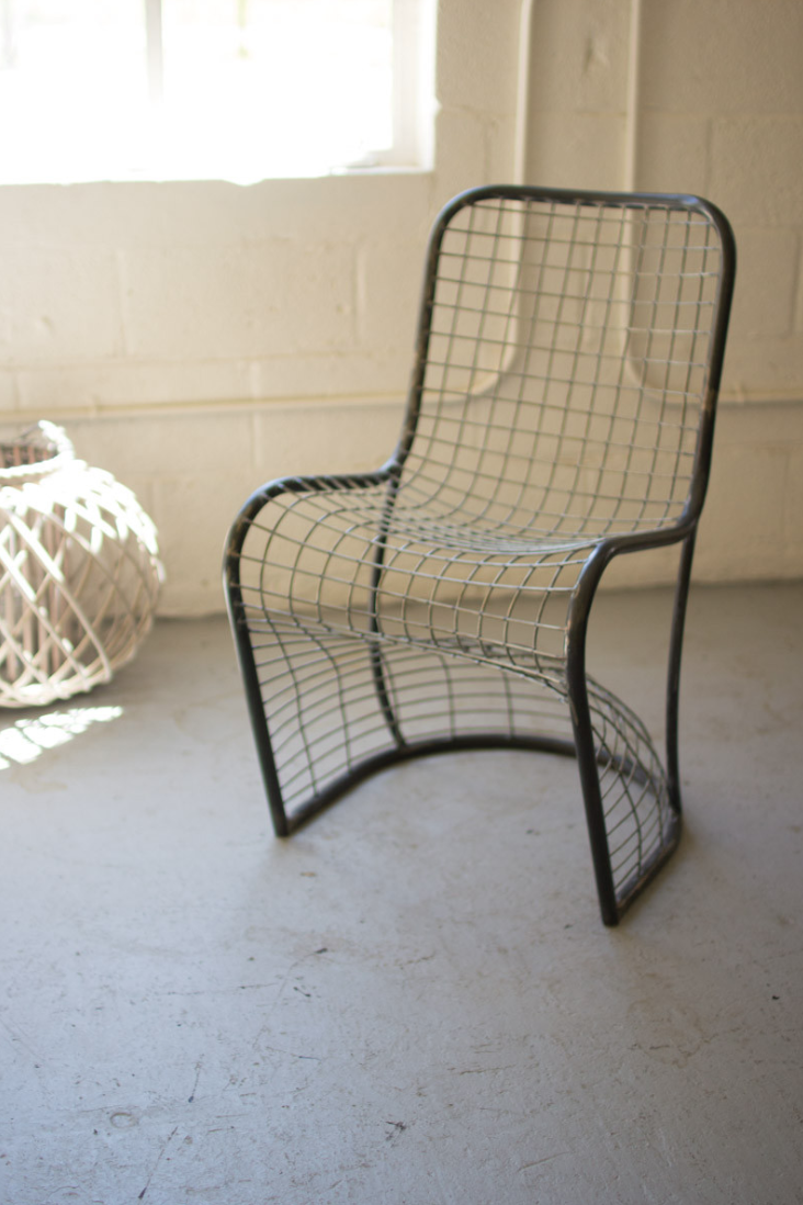 A Woven Metal S Chair from Kalalou, Inc with a black frame sits on a textured white floor in a sunlit bungalow with white walls and a decorative wicker object nearby.
