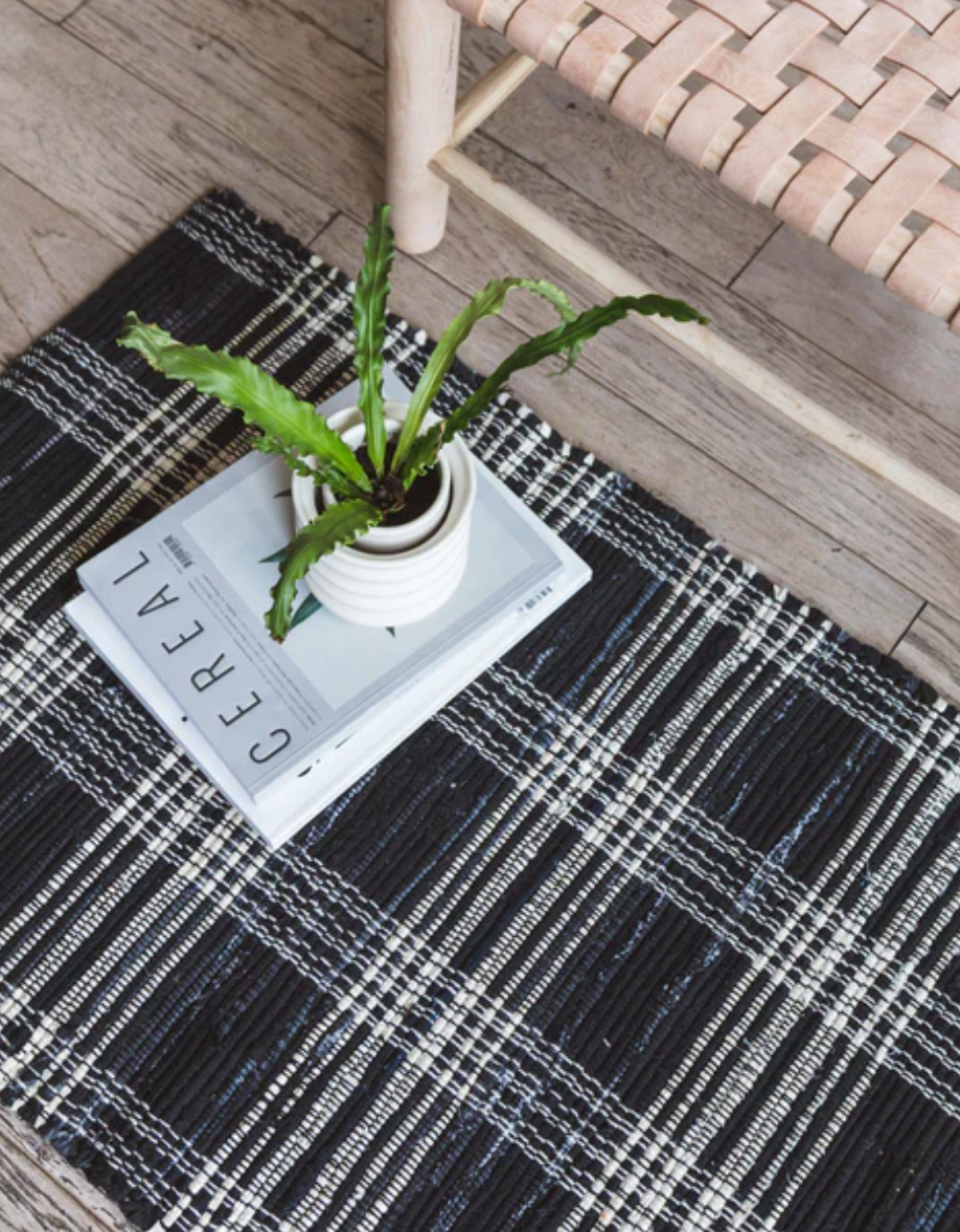 A small potted plant sits on top of two stacked magazines titled "Cereal," placed on a Plaid Black Denim Rug from Home Of The Brave. Part of a woven wooden chair is visible in the top right corner, all set on a wooden floor.