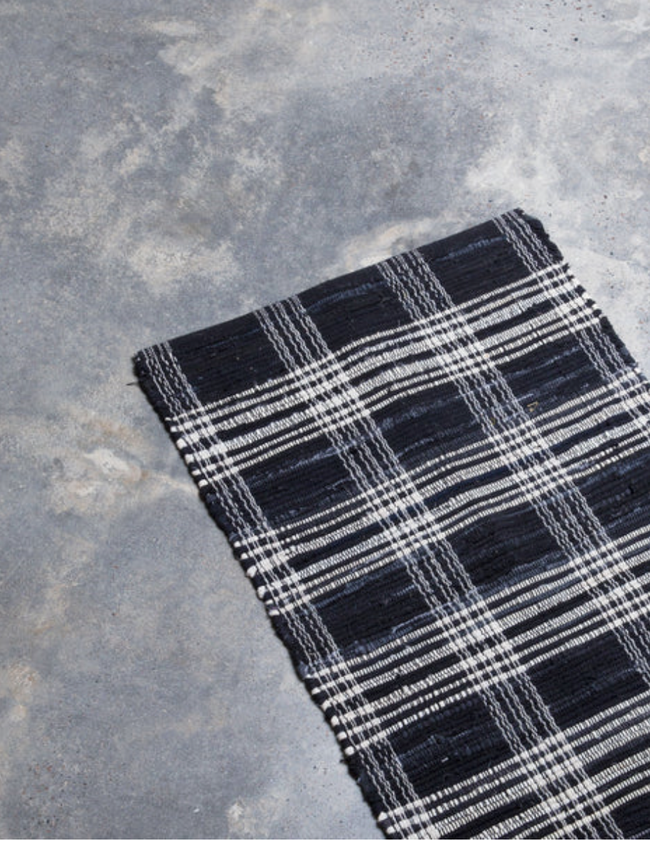 A Home Of The Brave Plaid Black Denim Rug is partially spread on a grey concrete floor. The rug's edge is visible at the bottom right corner of the image, featuring horizontal and vertical stripes that create a classic plaid design.