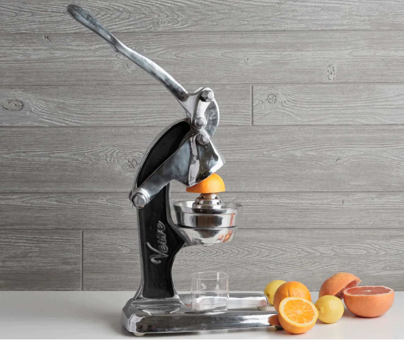 A Faire Mexican Citrus Juicer, crafted from recycled aluminum, sits on the counter with oranges and lemons. It's pressing an orange, ready to fill a glass against a gray wooden backdrop.