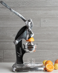 A Faire Mexican Citrus Juicer, crafted from recycled aluminum, sits on the counter with oranges and lemons. It's pressing an orange, ready to fill a glass against a gray wooden backdrop.